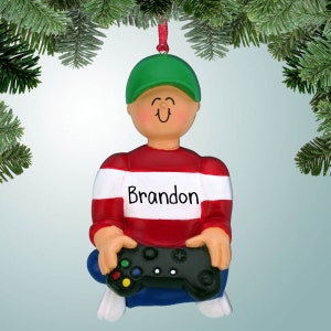 May include: A personalized Christmas ornament featuring a cartoon boy wearing a red and white striped sweater, a green hat, and blue pants. He is holding a black video game controller. The ornament is personalized with the name "Brandon".