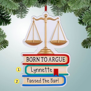 Può includere: Un ornamento a forma di bilancia della giustizia dorata con una pila di libri che recita "Born to Argue" e "Lynnette Passed the Bar!"