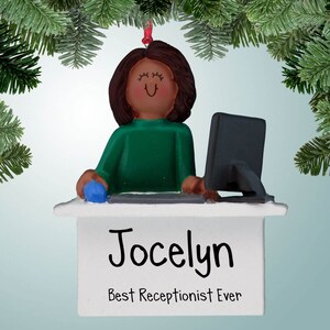 Puede incluir: Un adorno navideño personalizado con una mujer de dibujos animados de pelo castaño y un jersey verde sentada en un escritorio con un ordenador. El adorno tiene un escritorio blanco con un cartel que dice "Jocelyn La mejor recepcionista de todos los tiempos".