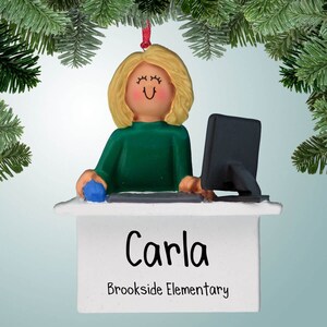 Puede incluir: Un adorno navideño personalizado que representa a una mujer de dibujos animados con cabello rubio sentada en un escritorio con una computadora. El adorno es blanco con un escritorio verde y un monitor de computadora negro. La mujer lleva un suéter verde. El adorno tiene el nombre "Carla" y el texto "Brookside Elementary" escrito en él.