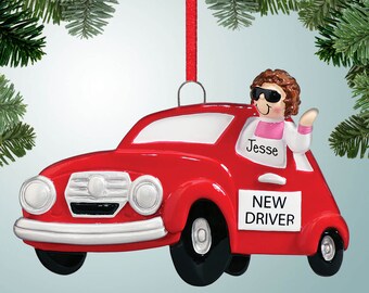 May include: Red car Christmas ornament with a person wearing sunglasses and a pink shirt driving the car. The car has a "NEW DRIVER" sign on the side and the name "Jesse" written on the window.