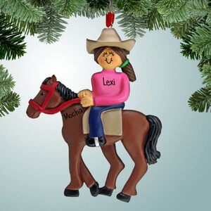 May include: A brown horse ornament with a black mane and tail, wearing a red bridle. A girl ornament is riding the horse, wearing a pink shirt, blue jeans, and a brown cowboy hat. The girl's name is "Lexi" and the horse's name is "Mocha".
