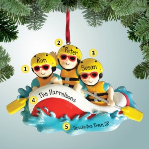 Puede incluir: Un adorno navideño con una familia de tres personas en una balsa amarilla en un río azul. La balsa está etiquetada como "The Harrelsons" y el río como "Deschutes River, OR". El adorno está decorado con colores rojo, amarillo y azul.