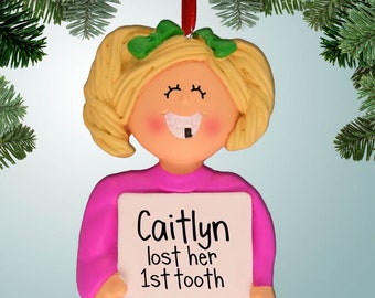Può includere: Un ornamento natalizio raffigurante una bambina sorridente con capelli biondi e fiocchi verdi. Indossa una maglietta rosa e tiene in mano un cartello con la scritta "Caitlyn lost her 1st tooth". L'ornamento è appeso a un nastro rosso.