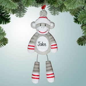 May include: A gray sock monkey Christmas ornament with red and white stripes. The monkey is wearing a white hat with a red pom-pom and has a white circle on its chest with the name "Jake" written in black.