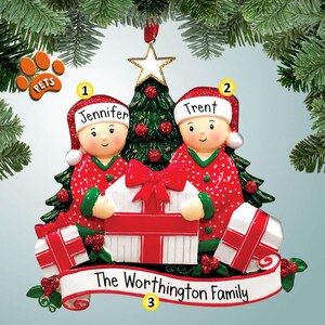 May include: A Christmas ornament featuring two figures in red pajamas holding a gift. The ornament includes a Christmas tree, star, and the names "Jennifer" and "Trent". The banner reads "The Worthington Family". A paw print with the word "PETS" is also included.