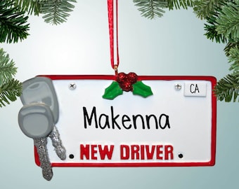 May include: A white and red Christmas ornament shaped like a license plate with the text "Makenna NEW DRIVER" and a pair of silver keys on the left side. The ornament has a red ribbon for hanging.