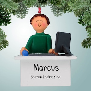 Puede incluir: Un adorno navideño personalizado que presenta a un hombre de dibujos animados con cabello castaño que lleva un suéter verde y está sentado en un escritorio con una computadora. El escritorio tiene una superficie blanca con el nombre "Marcus" y el texto "Search Engine King" escrito en él.