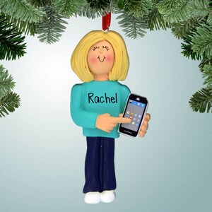 May include: Personalized Christmas ornament featuring a woman with blonde hair wearing a teal blue shirt and blue jeans. She is holding a smartphone and pointing at the screen. The ornament is personalized with the name "Rachel".