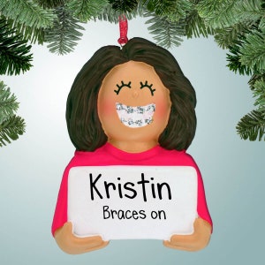 May include: A personalised Christmas bauble featuring a cartoon girl with brown hair and a pink shirt. She is smiling and wearing braces. The bauble has a white sign that says "Kristin Braces on".