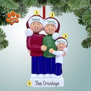May include: A personalised Christmas bauble featuring a family of three. The bauble is made of white ceramic and depicts a man, woman, and child wearing red and white Santa hats. The man is wearing a red sweater and blue jeans, the woman is wearing a green sweater and blue jeans, and the child is wearing a white sweater and blue jeans. The bauble is decorated with white snow and the family's last name, "The Crosbys", is written on the base.