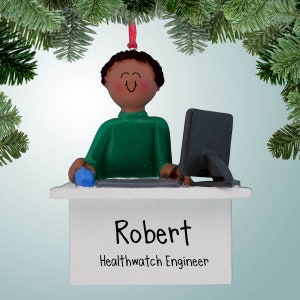 Puede incluir: Un adorno navideño personalizado que presenta una ilustración de dibujos animados de una persona con piel morena sentada en un escritorio con una computadora. El adorno es blanco con el nombre "Robert" y el título "Healthwatch Engineer" escrito en negro.