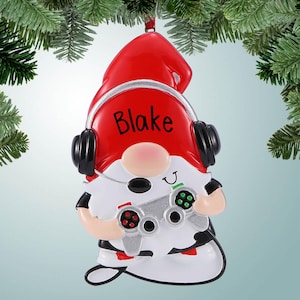 May include: A red and white gnome Christmas ornament with a black headset and a video game controller. The gnome is wearing a black and white shirt and has the name "Blake" written on its hat.
