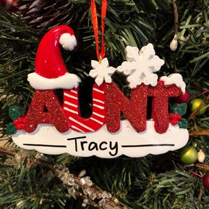 K&ouml;nnte beinhalten: Roter und wei&szlig;er Weihnachtsbaumschmuck mit dem Wort "AUNT" in glitzernden roten Buchstaben mit einem Weihnachtsmann-Hut auf dem "A". Der Schmuck hat eine wei&szlig;e Schneeflocke und den Namen "Tracy" auf einem wei&szlig;en Banner.