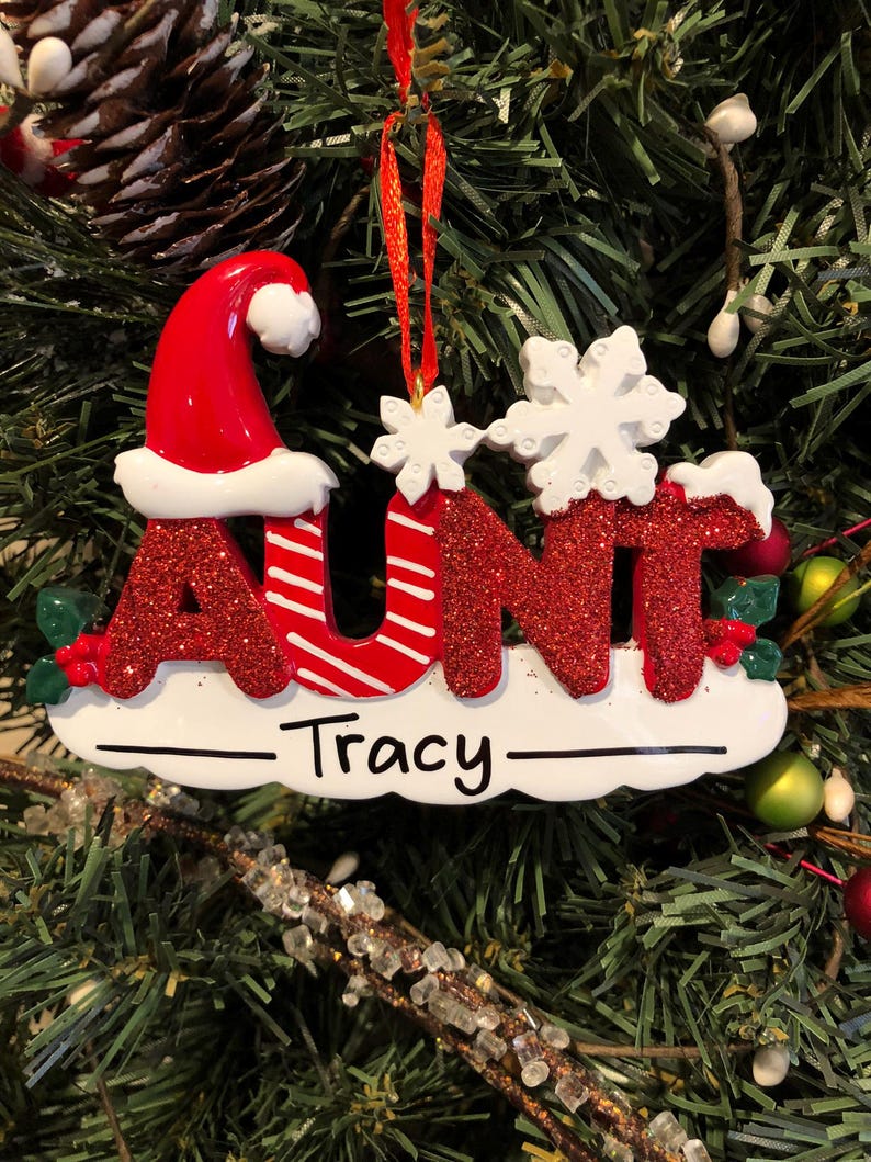 K&ouml;nnte beinhalten: Roter und wei&szlig;er Weihnachtsbaumschmuck mit dem Wort "AUNT" in glitzernden roten Buchstaben mit einem Weihnachtsmann-Hut auf dem "A". Der Schmuck hat eine wei&szlig;e Schneeflocke und den Namen "Tracy" auf einem wei&szlig;en Banner.