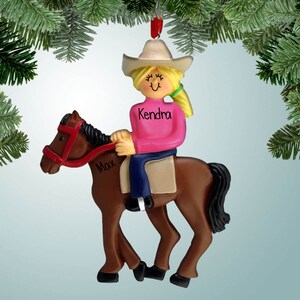 May include: A brown horse ornament with a girl riding it. The girl is wearing a pink shirt, blue jeans, and a cowboy hat. The horse has a red bridle and the girl's name, Kendra, is written on her shirt. The horse's name, Max, is written on the saddle.