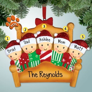 May include: A Christmas ornament depicting a family of five in a bed, wearing pajamas and Santa hats. The ornament is decorated with glitter and has the text "The Reynolds" on the bed.