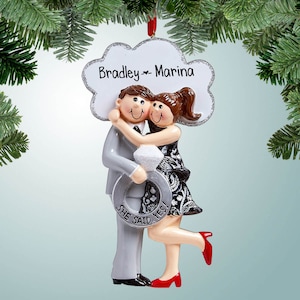 May include: A white and silver Christmas ornament featuring a cartoon couple embracing. The man is wearing a gray suit and the woman is wearing a black and white dress. The ornament has the text "Bradley & Marina" and "She Said Yes!" on it.