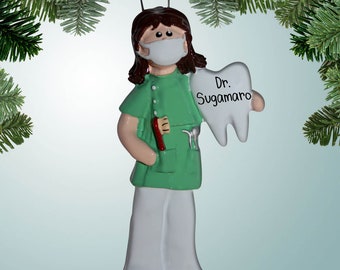 May include: A green and white Christmas ornament depicting a female dentist wearing a face mask and holding a large white tooth with the text "Dr. Sugamaro" on it.