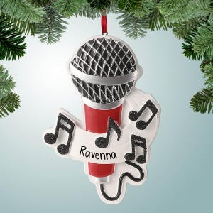 May include: A Christmas ornament shaped like a microphone, with a red base and a black and silver top. The ornament has a white banner with the name "Ravenna" and musical notes. It hangs from a red ribbon.