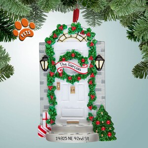 May include: A white door with a green holly wreath and red berries. The door has a sign that says "The Garcias" and a small Christmas tree in front of it. The address "14825 NE 42nd St" is written below the door. This is a personalized Christmas ornament.