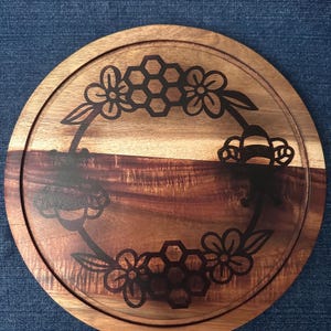 May include: Round wooden serving board with a decorative floral and honeycomb design. The board is made of a light brown wood with a darker brown wood grain pattern. The design is etched into the wood, creating a contrasting dark brown color.