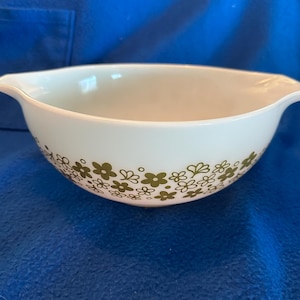 May include: White ceramic bowl with a floral pattern in olive green. The bowl has a curved shape with handles on either side. The floral design is repeated around the bowl's exterior.