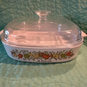 May include: Oval white casserole dish with a clear glass lid and handle. The dish features a decorative band of colorful fruits and vegetables, with the text "Le Romarin" below the design.