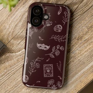 May include: A dark maroon smartphone case featuring a white line art pattern of various botanical elements, a masquerade mask, a ring, feathers, and a small rectangular card with the text Witch Magic printed in a thin, delicate font style.