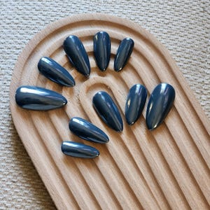 May include: A set of ten glossy, dark teal-colored false nails arranged on a light brown, wooden, arched tray. The nails have a pointed, almond shape and a metallic finish, reflecting light. The tray has a ridged design.