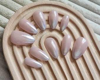 May include: A set of ten oval-shaped, light pink false nails with an iridescent shimmer effect. The nails are arranged on a light brown wooden tray with a grooved design. The nails are a beauty product.