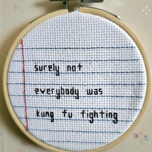 Op de afbeelding: Een witte borduurring met een kruissteekontwerp met de tekst "zeker niet iedereen was kung fu aan het vechten".