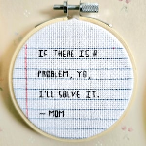 May include: A white embroidery hoop with a blue and white grid pattern. The text "IF THERE IS A PROBLEM, YO. I'LL SOLVE IT. -- MOM" is embroidered in black thread.