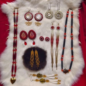 Peut inclure: Une collection de bijoux artisanaux comprenant deux longs colliers de perles, l'un avec des perles cylindriques rouges et un pendentif cœur, l'autre avec des perles noires et rouges. Plusieurs paires de boucles d'oreilles perlées aux tons rouge, or et blanc.