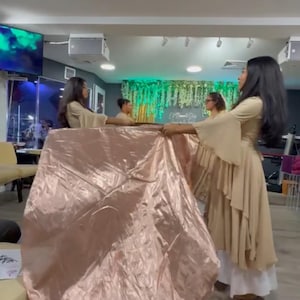 May include: Several people are holding up a large, shiny, pink fabric. They are wearing long, flowing, beige dresses. The scene appears to be indoors, possibly a performance or rehearsal space. The lighting is bright, and the overall aesthetic is elegant.
