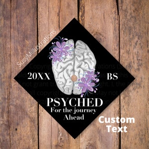 Op de afbeelding: Zwarte afstudeerhoed met een witte hersenenafbeelding en paarse bloemen. De tekst "PSYCHED For the journey Ahead" is op de hoed gedrukt. De tekst "Custom Text" is onder de hoed gedrukt.
