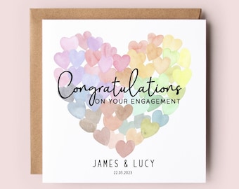 May include: A white card with a watercolor heart made of many small hearts in shades of pink, blue, yellow, and green. The text "Congratulations on your engagement" is written in black script font. Below the heart, the names "James & Lucy" and the date "22.05.2023" are printed in black.