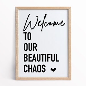 May include: A framed print with the text "Welcome to our beautiful chaos" in black lettering with a small heart symbol at the end. The print is in a light wood frame.