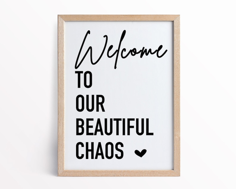May include: A framed print with the text "Welcome to our beautiful chaos" in black lettering with a small heart symbol at the end. The print is in a light wood frame.