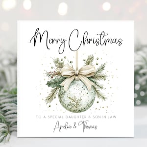 May include: A square Christmas card with the words "Merry Christmas" in elegant script. The card features a watercolor illustration of a Christmas ornament with a bow, greenery, and the text "TO A SPECIAL DAUGHTER & SON IN LAW" and the names "Amelia & Thomas".
