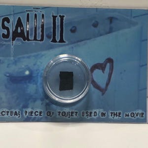 Könnte beinhalten: Eine transparente Kunststoffhülle mit einem schwarzen Quadrat, vor blauem Hintergrund mit dem Text "SAW II" und "ACTUAL PIECE OF TOILET USED IN THE MOVIE". Ein rotes Herz ist auf dem Hintergrund gezeichnet.