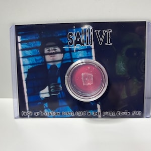 May include: A collectible card from the film "Saw VI" featuring a piece of imitation glass. The card is encased in clear plastic, with a background image of a man in a suit and a green-faced character. The text "SAW VI" is visible.