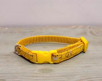 Puede incluir: Un collar amarillo para gato con una hebilla de plástico amarilla a juego y una anilla en D de metal plateado. El collar está hecho de un material tejido amarillo con un patrón repetido de pequeñas formas negras y blancas.