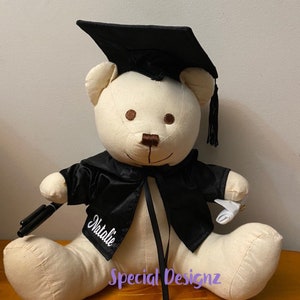 May include: A white plush teddy bear wearing a black graduation gown and cap. The bear is holding a diploma and a pen. The bear is personalised with the name "Natalie" and the text "Special Designz".