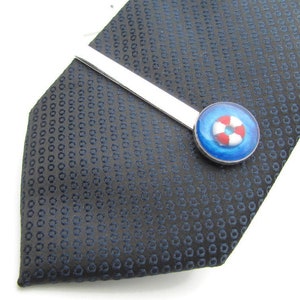 Peut inclure: Un fermo à cravate argenté avec un charme de bouée de sauvetage en émail bleu et blanc. Le fermoir à cravate est sur une cravate à motif bleu foncé.
