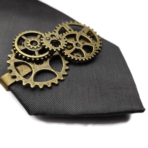 May include: A gold-toned tie clip featuring a design of interlocking gears. The clip is attached to a dark grey tie.