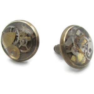 Op de afbeelding: Een paar gouden oorbellen met een rond, transparant harsontwerp met een steampunk-geïnspireerd uurwerkpatroon.