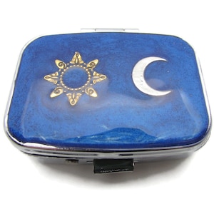 May include: A small, blue metal compact with a gold sun and a silver crescent moon design.