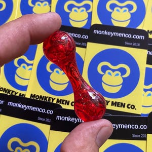 Op de afbeelding: Een rood, zandloper-vormig object met zwevende glitter, vastgehouden tussen vingers. De achtergrond toont gele en blauwe kaarten met een apenlogo en de tekst "monkeymenco.com Since 2018".