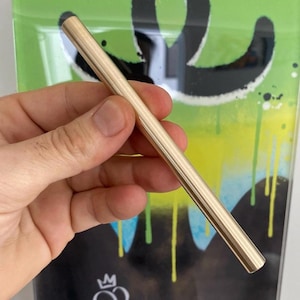 Puede incluir: Un lápiz óptico dorado, de forma cilíndrica, sostenido contra un fondo de arte graffiti verde y negro. El fondo presenta un diseño de estilo graffiti con goteos y salpicaduras. El lápiz óptico parece ser de metal.